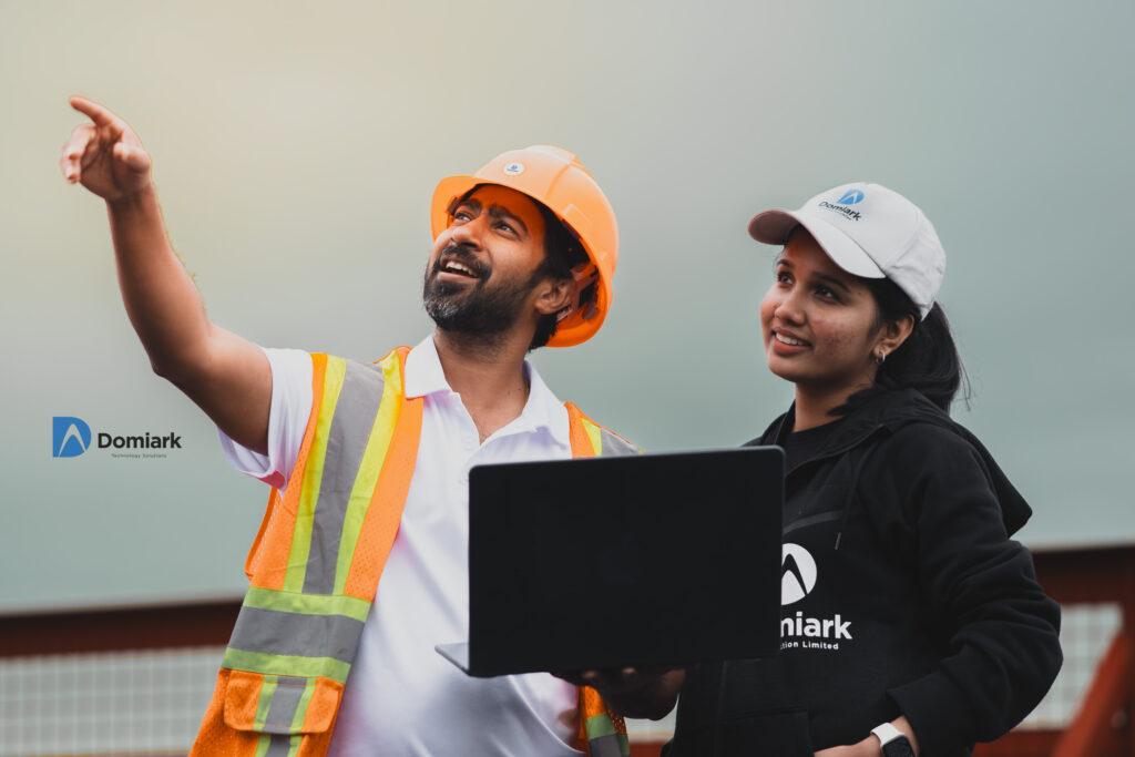 A male site manager in an orange hard hat and safety vest points off-camera while holding a laptop and speaking with a female colleague wearing a Domiark baseball cap and black hoodie outdoors near structural equipment.
